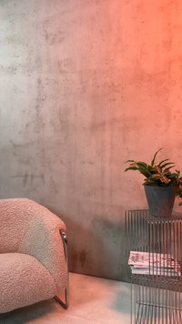 Gebogener beiger Bouclé-Stuhl neben einem Metall-Draht-Beistelltisch mit Blumentopf und gestapelten Zeitschriften vor einer strukturierten Betonwand.