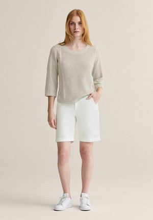 Kvinde med rødt hår iført en beige strikket sweater, hvide knælange shorts og hvide sneakers, stående med den ene hånd i lommen.