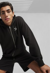 Sort zip-up hoodie med en frontlomme, snørebånd og Puma-logo på venstre bryst. Båret med matchende sorte shorts.
