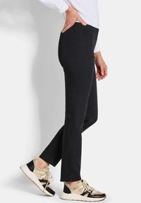 Vrouw in zwarte slim-fit broek, witte lange mouwen shirt en beige-zwarte sneakers, staande met één hand op haar heup.