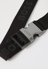 Crni platneni remen s utisnutim "BOSS" logom, sa metalnom srebrnom kopčom i glatkom teksturom, prikladan za svakodnevno ili formalno nošenje.