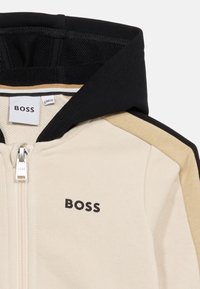 Μπεζ και μαύρο φούτερ με φερμουάρ, με μαύρο λογότυπο "BOSS" στο στήθος, μανίκια σε μπλοκ χρωμάτων μ πεζ και μαύρου, και μαύρη εσωτερική επένδυση κουκούλας.
