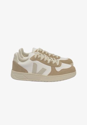 Par de zapatillas bajas beige y blancas con paneles de ante y piel perforada, cordones beige y un logo "V" beige en el lateral.