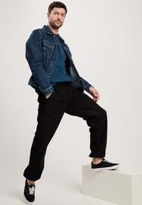 Jeansowa kurtka, turkusowa koszulka polo i czarne spodnie cargo. Czarne sneakersy z białymi podeszwami. Model pozuje na białym sześcianie na tle jasnego tła.