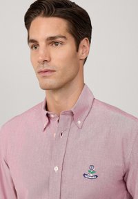 Hombre con cabello corto castaño que lleva una camisa de botones rosa clara con un pequeño logo bordado en el pecho, mirando ligeramente hacia la izquierda.