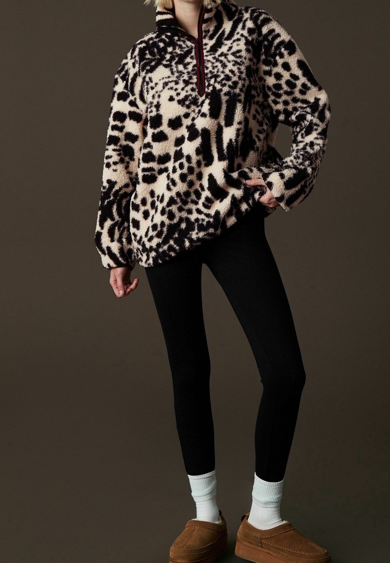 Osoba ubrana w czarno-biały polar z motywem zwierzęcym, czarne legginsy, białe skarpetki i brązowe buty wsuwane na ciemnym tle.