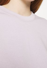 Mujer con cabello rojo que lleva una camiseta de cuello redondo de color púrpura claro, mostrando el escote y la parte superior del pecho contra un fondo blanco.