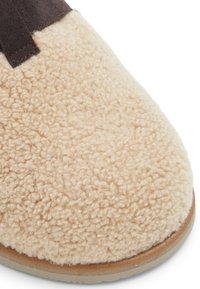 Beige schapenleren pantoffel met een zacht textuur bovenwerk en bruine suède accenten. Ronde neus, rubberen zool en gestikte details.