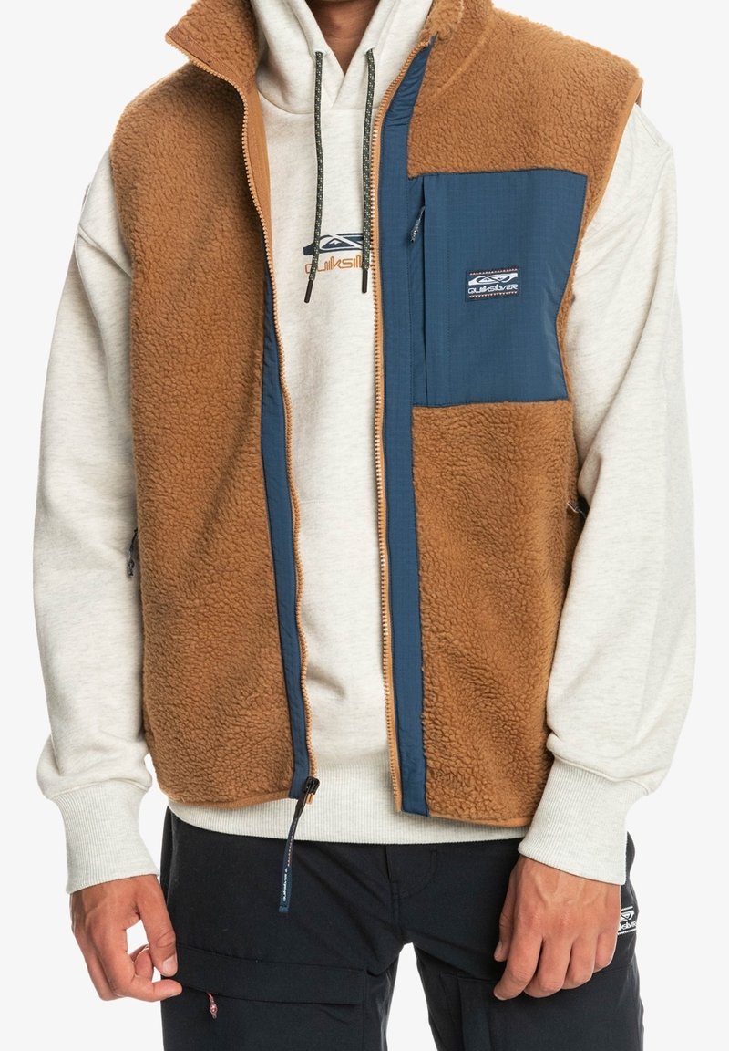 Brun fleeceväst med blå nylonfrontpanel, har en dragkedja, bröstficka och sidfickor över en grå huvtröja och svarta byxor.