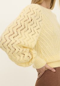 Żółty sweter z dzianiny o fakturze zygzakowatego wzoru, z bufiastymi rękawami i ściągaczami na mankietach. Miękki, lekki materiał, luźny krój.