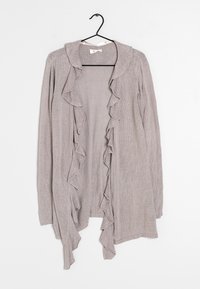 Vaaleanharmaa neuletakki, joka on valmistettu teksturoidusta neulosmateriaalista ja jossa on röyhelötrimmi etureunoilla ja kaula-aukossa. Ripustettuna mustasta koukusta.