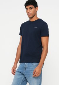 Σκούρο μπλε βαμβακερό μπλουζάκι με κοντά μανίκια, στρογγυλή λαιμόκοψη και μικρή λεπτομέρεια λογοτύπου στο αριστερό στήθος. Συνδυασμένο με ανοιχτό μπλε τζιν.