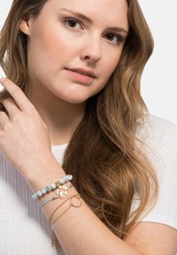 Una pila di bracciali con un charm a forma di globo dorato, perline bianche, un bracciale azzurro chiaro e una catena dorata. La modella mostra gli accessori sul suo polso.