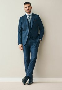 Dunkelblauer Anzug mit einem Blazer mit drei Knöpfen, passender Weste und Hose. Kombiniert mit einer gemusterten Krawatte und schwarzen Lederschuhen. Strukturiertes Material.