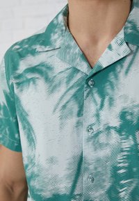 Camisa de manga corta y cuello en tonos verde azulado y azul claro con patrones ondulados; presenta dos botones y una textura suave y lisa.