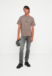 Man wearing gray Calvin Klein t-shirt, faded gray jeans, black shoes, holding small gray bag against plain white background.