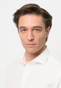 Un primo piano di un uomo che indossa una camicia bianca semplice con colletto alla canadese e una leggera texture, su uno sfondo neutro.