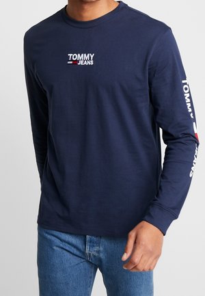 Uomo che indossa una maglia a maniche lunghe blu navy "Tommy Jeans" con logo sul petto e sulla manica, abbinata a jeans blu, su sfondo semplice.