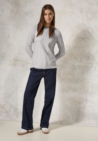Maglione grigio chiaro a trecce con scollo rotondo, abbinato a jeans blu scuro dalle linee morbide e sneaker grigie e bianche. Sfondo neutro.