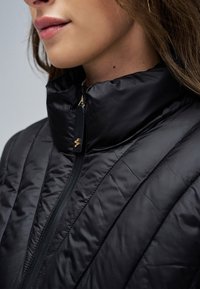 Nahaufnahme einer Frau, die eine schwarze gesteppte Jacke mit hohem Kragen und einem Reißverschluss mit kleinem goldenem Zipper-Anhänger trägt.