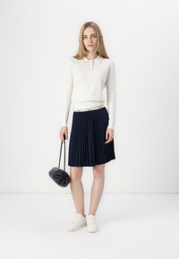 White ribbed long-sleeve polo, navy pleated skirt, black handbag, and white sneakers. Smooth textures, fitted design, and contrasting colors.