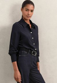 Navy button-up shirt with a structured collar, long sleeves, and gather at cuffs, paired with dark jeans and a black belt with silver hardware.