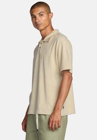 Beige polo tee med et subtilt tekstureret mønster, to-knaps lukning og korte ærmer. Båret med lysegrønne bukser.