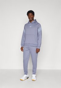 Felpa grigia chiara con cappuccio e tasca frontale, con branding blu sulle maniche, abbinata a pantaloni felpati coordinati e scarpe bianche.
