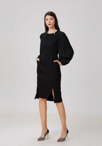 Robe noire en tissu texturé, avec des manches bouffantes, des poches latérales et des fentes montantes jusqu'à la cuisse. Fini avec des talons hauts pointus et élégants.