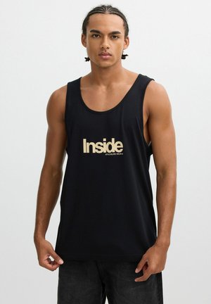 Joven con cabello trenzado que lleva una camiseta sin mangas negra con el texto "Inside Athleisure Wear", posando frente a un fondo blanco liso.