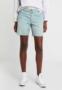 Personne portant un short vert clair, une ceinture blanche, un haut à manches longues gris et des baskets montantes blanches, se tenant devant un fond uni.