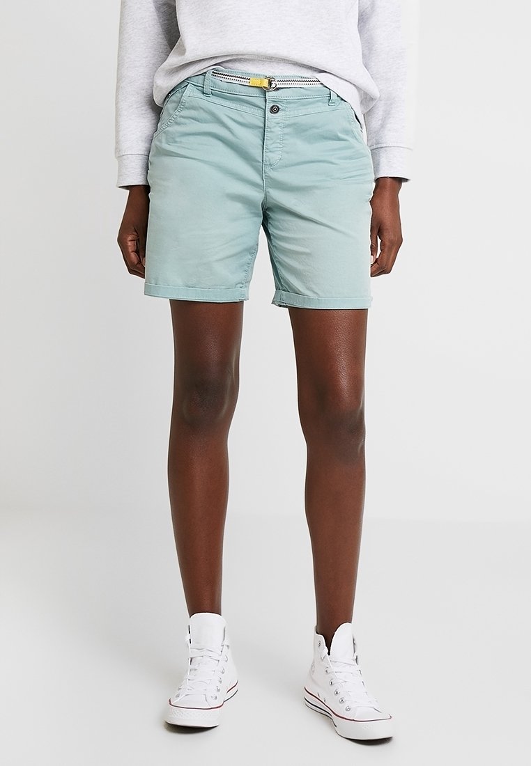 Personne portant un short vert clair, une ceinture blanche, un haut à manches longues gris et des baskets montantes blanches, se tenant devant un fond uni.