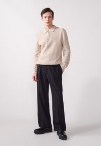 Pull en tricot beige avec une fermeture éclair et des côtes verticales, associé à un pantalon large noir et des chaussures noires épaisses.