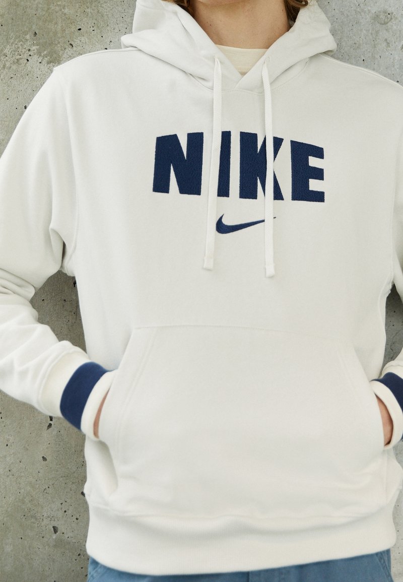 Vit huvtröja med en stor marinblå "NIKE" logotyp och en liten swoosh, med ribbad marinblå muddar och en känguruficka. Mjuk tygkänsla.