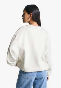 Una donna con lunghi capelli scuri è in piedi, di spalle, indossando una felpa bianca oversize e jeans azzurri contro uno sfondo bianco neutro.