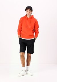 Orange hoodie with drawstrings and front pocket, paired with black shorts. Footwear is white sneakers, complemented by white socks.