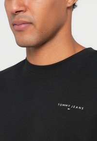 Μαύρο t-shirt με λαιμόκοψη, κατασκευασμένο από βαμβάκι, που διαθέτει το λευκό λογότυπο "TOMMY JEANS" στο αριστερό στήθος και μια μικρή λεπτομέρεια σε κόκκινο και μπλε χρώμα.