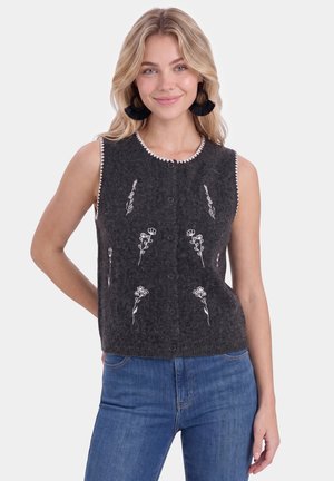 Blonde vrouw die een mouwloos donkergrijs vest met witte bloemkanten en een blauwe spijkerbroek draagt, glimlachend met één hand achter haar rug.