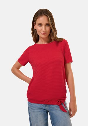 Haut court à manches courtes rouge texturé avec un col rond et un détail noué sur le côté, assorti à un jean en denim bleu clair.