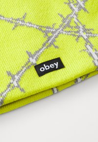 Žiarivo žltá pletená čiapka s šedobielym vzorom ostnatého drôtu. Obsahuje čierny štítok so slovom "obey". Mäkký, štruktúrovaný materiál.