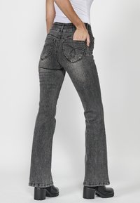 Jeans de mezclilla negros acampanados con una cintura de media altura, que presentan bolsillos traseros con costuras curvas y una textura suave. Combinados con botas de tacón.