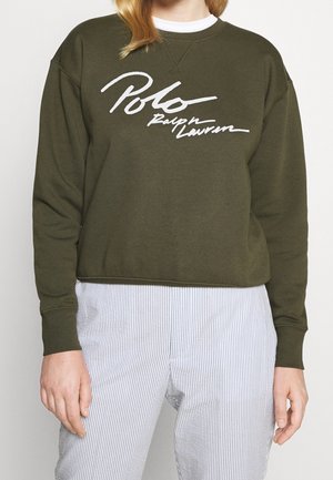 Person iført olivengrøn sweatshirt med hvid "Polo Ralph Lauren" tekst og lyse, lodretstribede bukser, stående mod en ensfarvet baggrund.