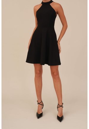 Kobieta ubrana w czarną sukienkę na ramiączkach z rozkloszowanym dołem oraz czarne, szpilki z noskiem w szpic i paskiem wokół kostki.