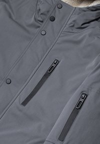 Grå jacka med mjukt inre, med svarta tryckknappar och dragkedjefickor fram, tillverkad av slätt, texturerat tyg.