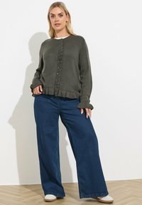 Ciemnozielony sweter z dzianiny z falbaniastym wykończeniem u dołu i na mankietach, z zapięciem na guziki, w połączeniu z szerokimi niebieskimi spodniami dżinsowymi. Gładka tekstura, swobodny styl.