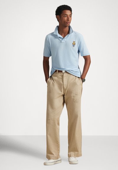 Fiatal férfi áll zsebében kezével, világoskék pólóingben, amin hímzett medve látható, bézs színű bő nadrágban és fehér tornacipőben.