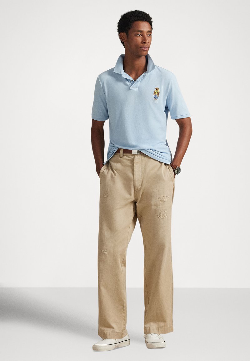 Junger Mann mit Händen in den Taschen, trägt ein hellblaues Poloshirt mit gesticktem Bärenmotiv, bequeme beige Hose und weiße Sneakers.