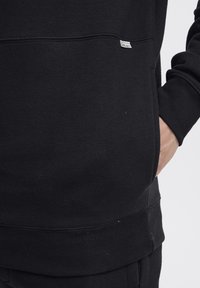 Sudadera negra con cuello redondo, puños y dobladillo acanalados, con una pequeña etiqueta blanca en el lateral. El material parece suave y texturizado.
