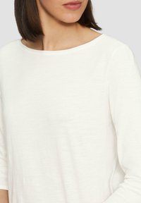 Haut blanc à manches longues avec un col rond, fabriqué en tissu léger. Présente une texture lisse et un minimum de détails de couture.