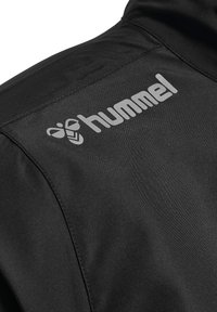 Svart lättviktsjacka med en slät textur, med en framträdande "hummel"-logotyp i grått på den övre vänstra sidan.
